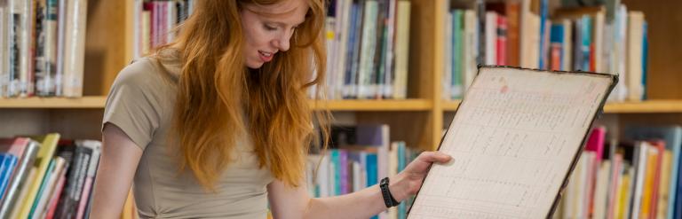 A woman looks through historical records with library shelves behind her