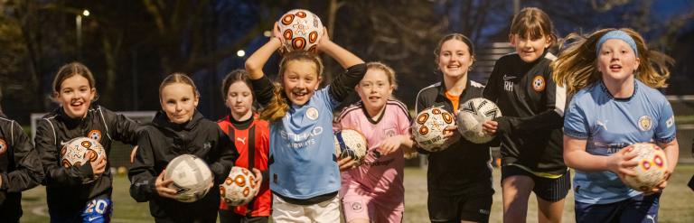 Eight girls running forwards in a line carrying footballs