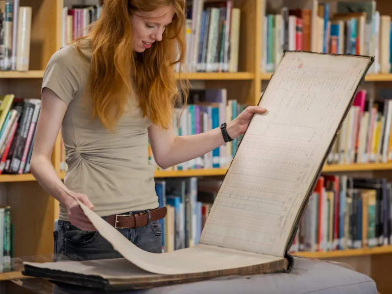 A woman looks through historical records with library shelves behind her