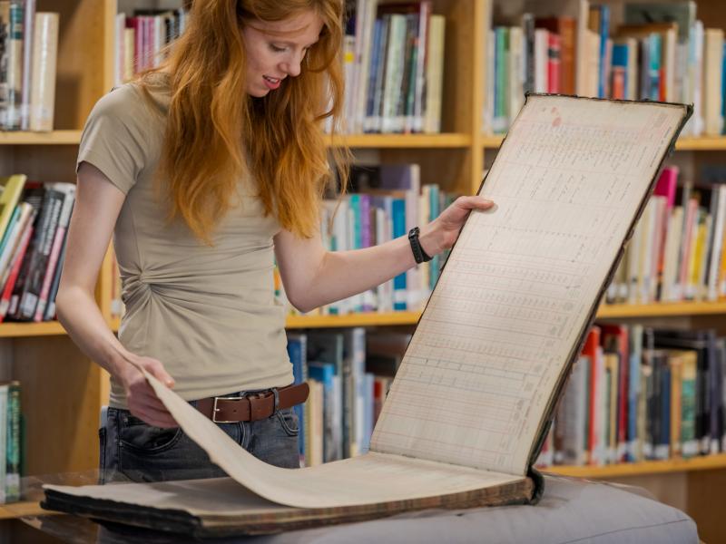 A woman looks through historical records with library shelves behind her