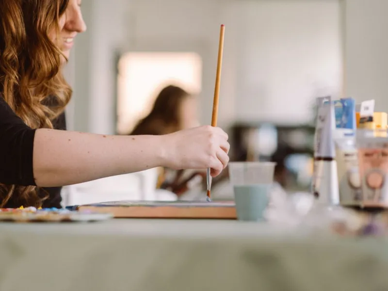 Side view of young women painting at a table in foreground with blurred background.