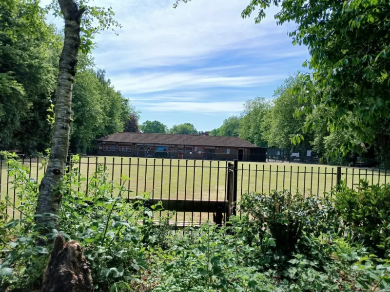 A bowling hut seen from across a bowling green with trees and greenery in front of railings in the foreground