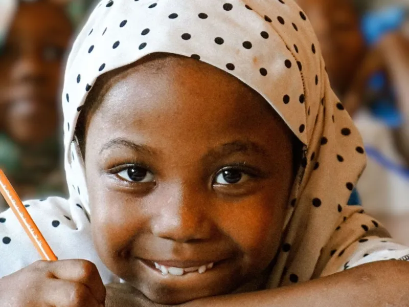 Child in hijab smiling and holding a pencil.
