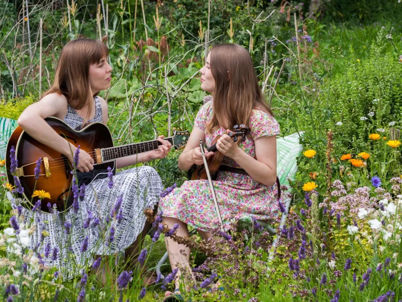 Two women in a meadow, singing together on guitar and ukelele