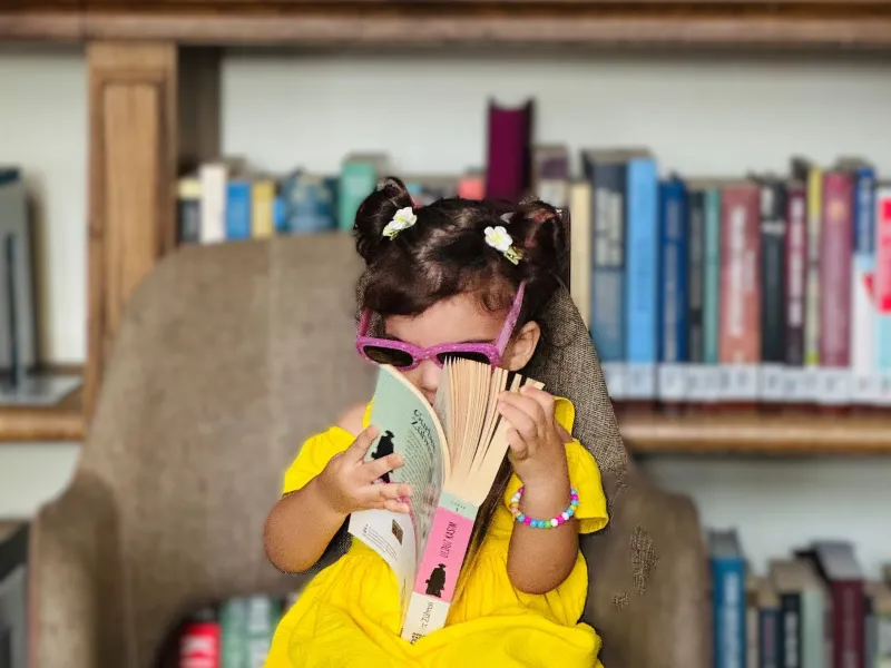 Little girl in sundress and sunglasses sat in big chair reading big book in a library setting.