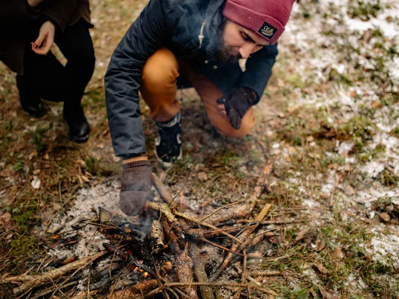 A man kneeling next to a small fire placing sticks on it