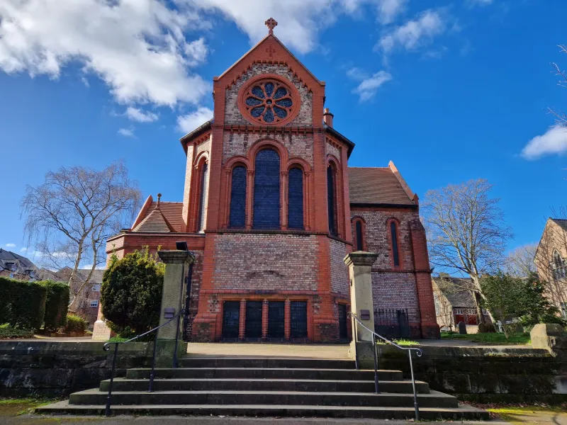 Exterior view of St George’s Church, Altrincham, a large historic red-brick church with tall arched windows and a distinctive circular rose window. Stone steps and railings lead up through the churchyard to the building, beneath a bright blue sky with scattered clouds.