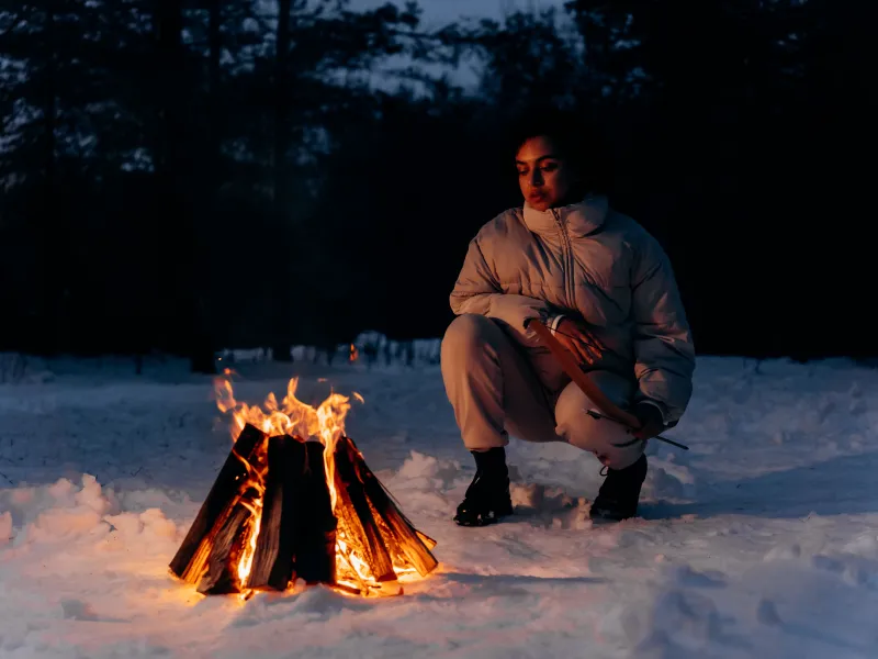 A woman crouching near a fire on snow covered ground with trees in the background