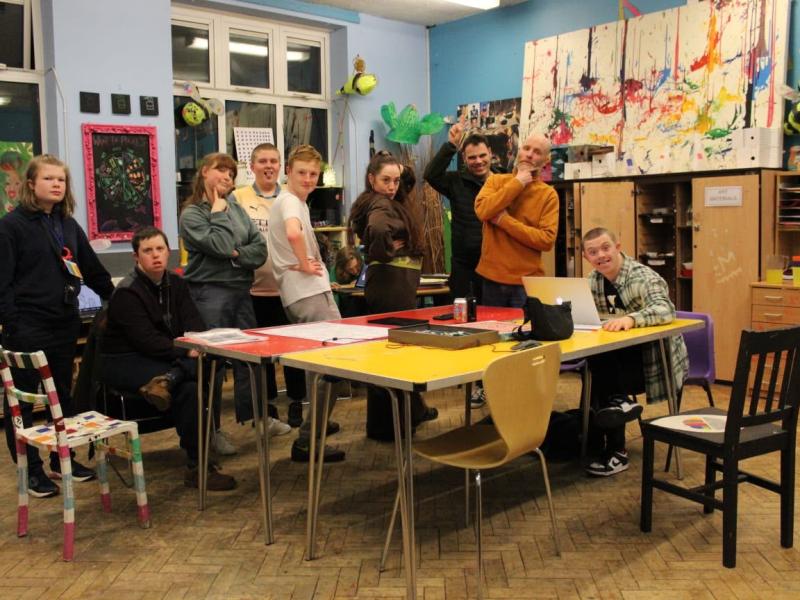 A group of young people and staff pose playfully around tables in the colourful art room at Gorse Hill Studios, surrounded by paintings, craft materials and decorated furniture.