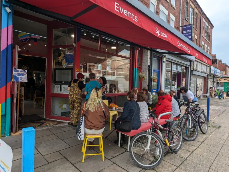 A view of OT Creative Space, with a red awning and people looking at artwork in the window from seats out front 