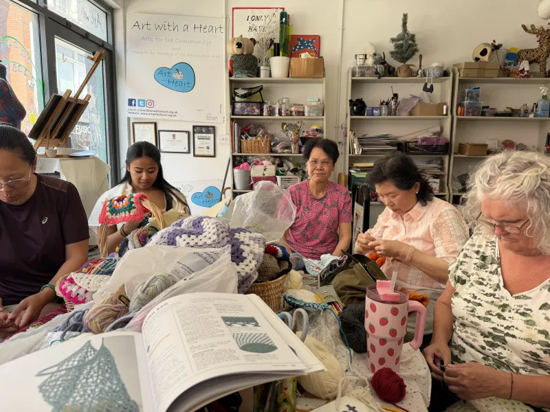 A group of women of different ages sit around a table at Art with a Heart, crocheting together among colourful yarn, handmade blankets and crochet books.