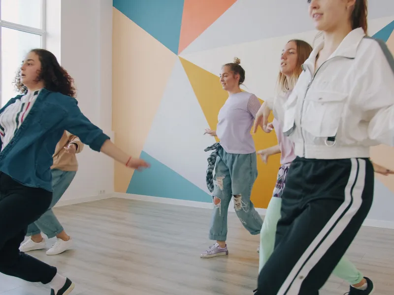 Five women dancing in a room with a wooden floor and multicoloured wall