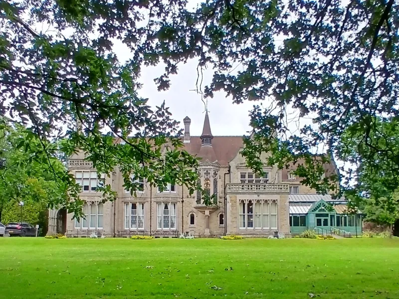 A view across a lawn in a park with foliage in the top of the frame, a grand house in the distance