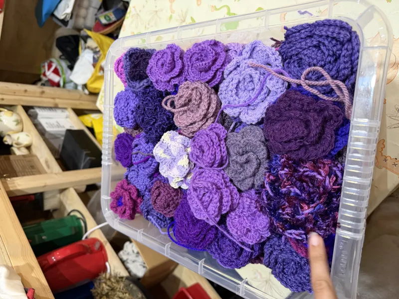 A storage box full of different shades of purple crochet roses sat on a craft table in art with a heart