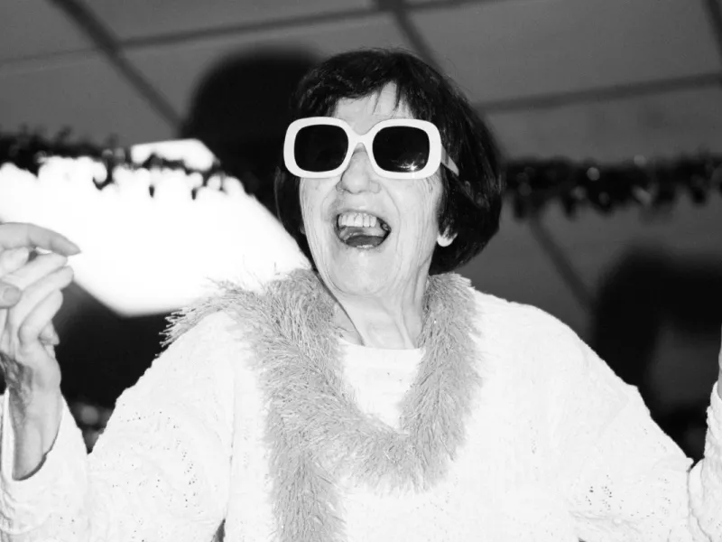 An older woman with large white-rimmed sunglasses is joyfully singing or shouting and holding hands with someone off-frame