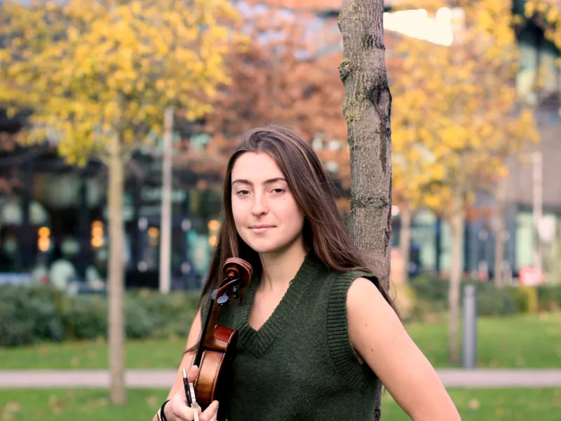 A photo of Esther standing in a park holding her violin under her right arm.
