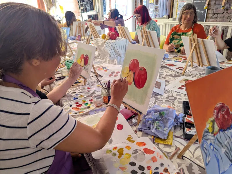 People sat around a table painting still life apples on small easels with oil paints 