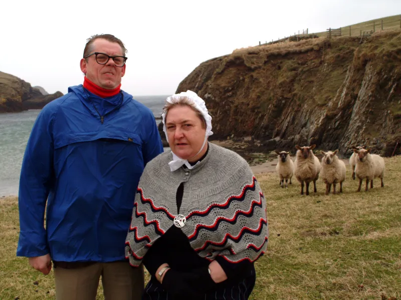 A man in a blue anorak and a woman in a grey shawl standing in front of some sheep