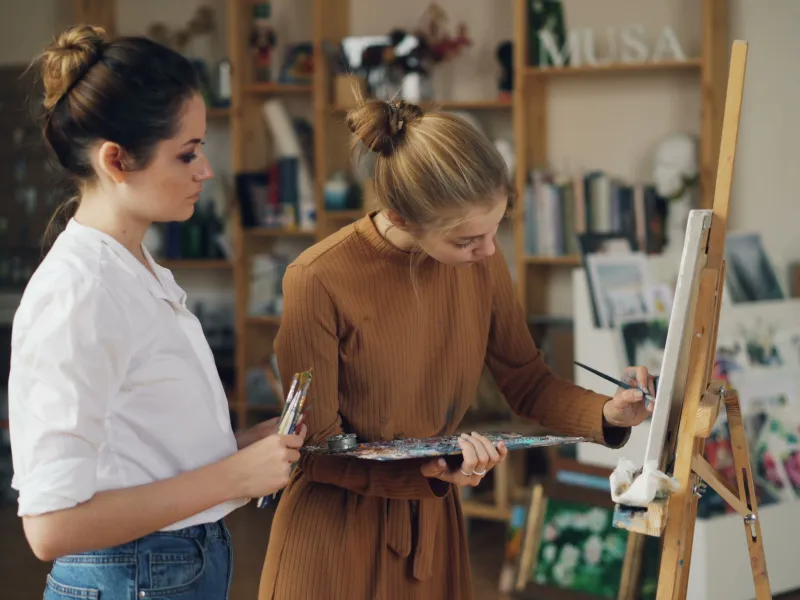 Two women stood next to an easel, one painting and the other watching 
