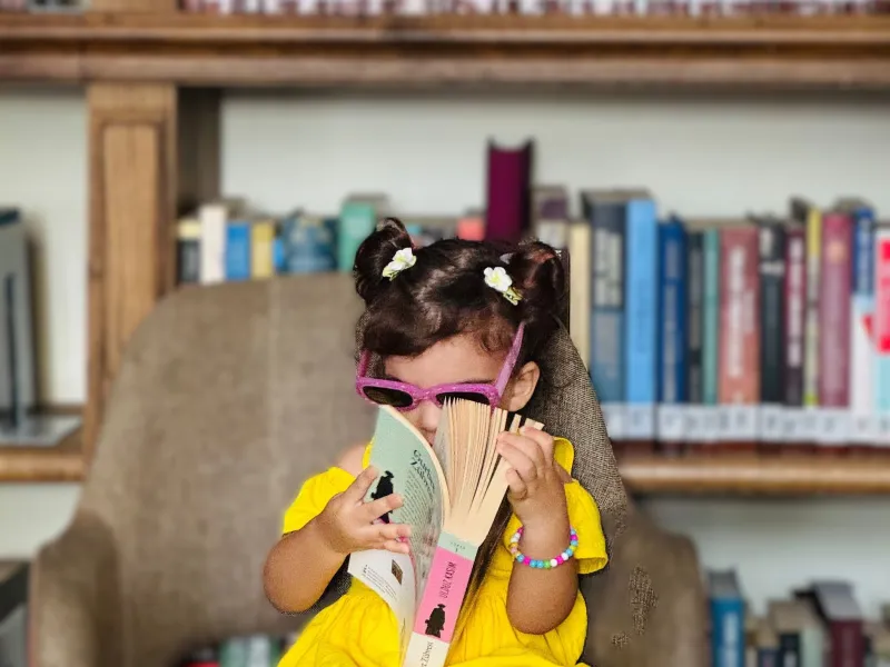 Little girl in yellow dress with sunglasses on sat on a big chair holding a very big book - with background of book shelves.