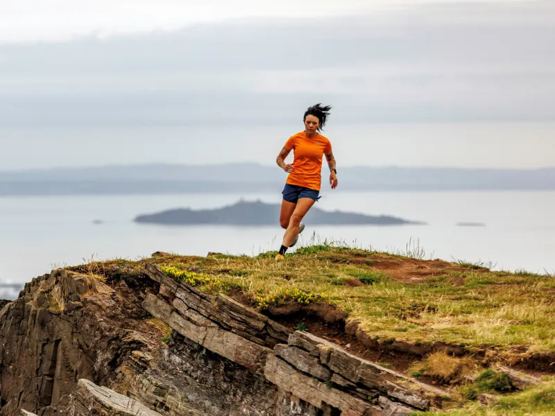 A woman in an orange top running along a cliff top