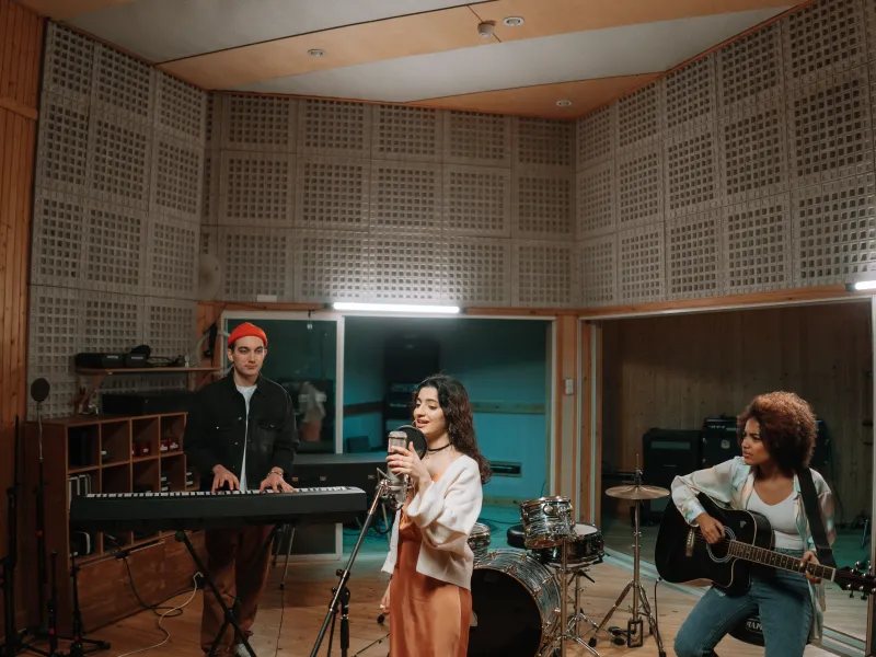 Three people stood in a music studio, one playing a keyboard, one playing a guitar and one singing into a microphone