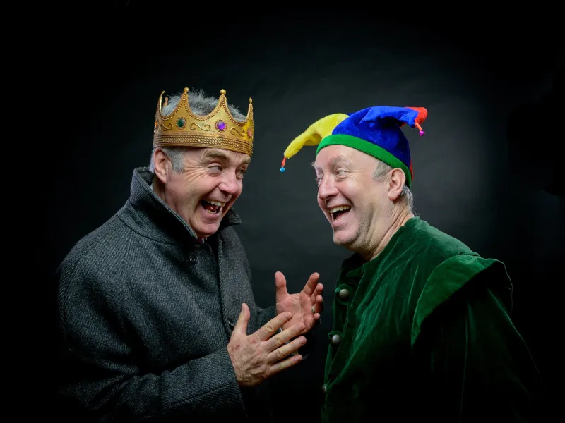 A man in a crown and a second in a jester's hat laughing at each other